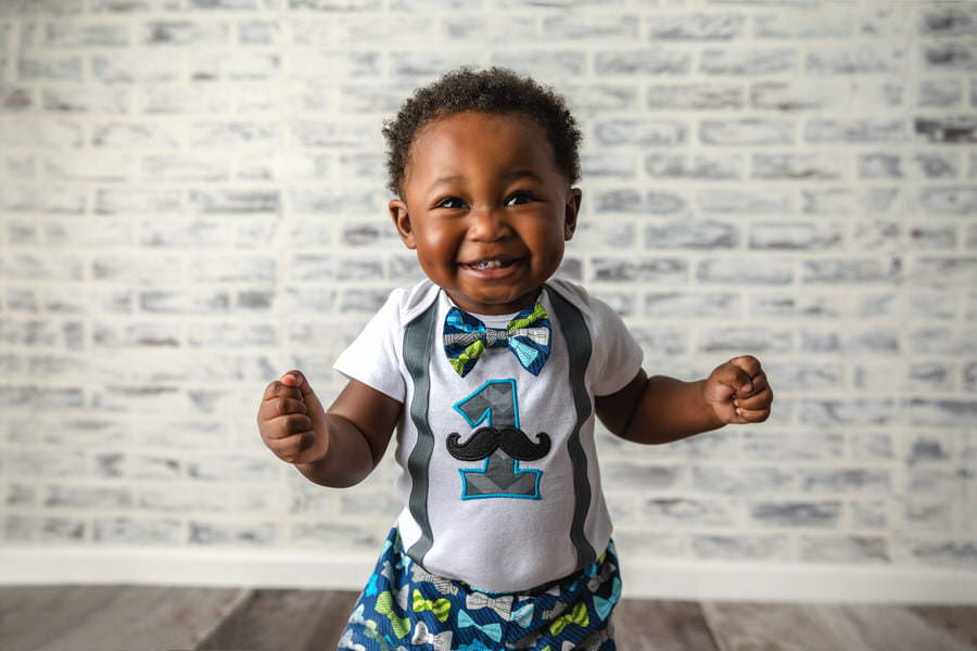 Excited black baby boy standing in front of a white brick wall in a blue and green mustache outfit during his cake smash photoshoot