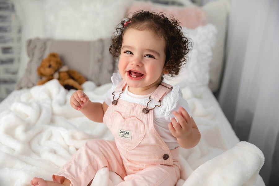Baby girl in pink overalls sitting on a bed laughing during her first birthday photoshoot