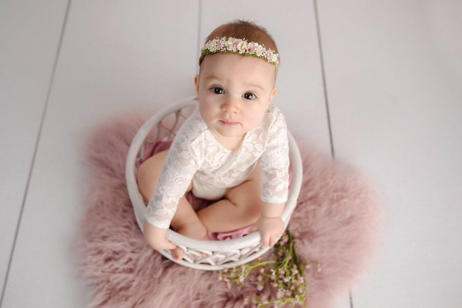 Baby girl in a lace outfit looking up at the camera sitting in a white prop during her baby photoshoot