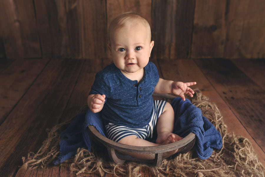 Smiling 6 month old baby boy in a blue outfit sitting in a brown bowl posing during his baby photoshoot