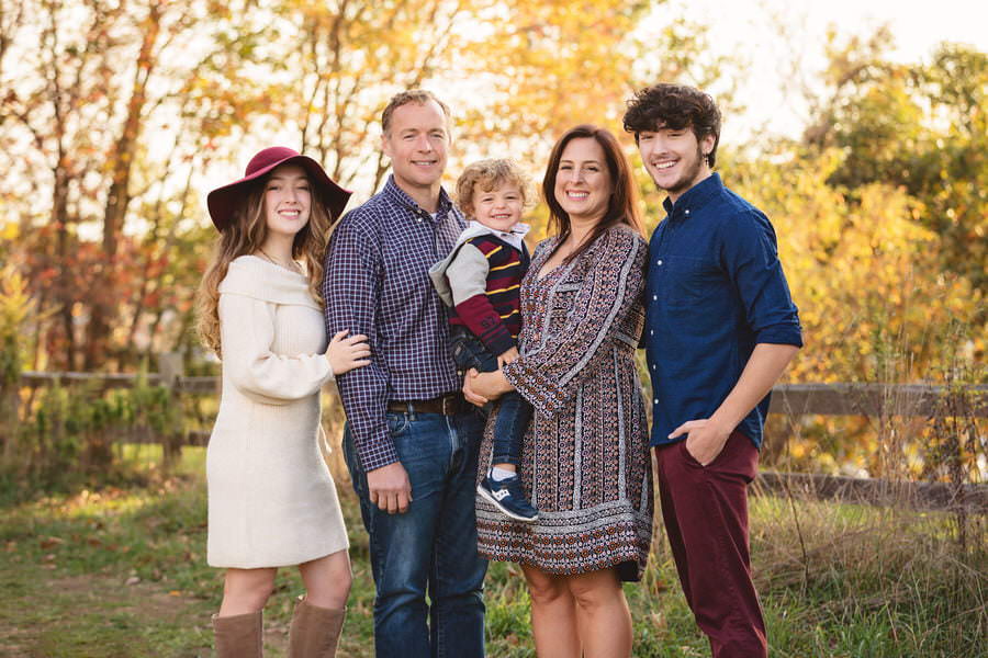 Family of 5 standing and smiling at the camera with yellow fall foliage behind them during their family photoshoot