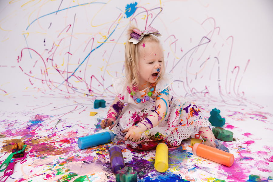 Expressive toddler girl playing with paint bottles with rainbow colors of paint behind her during her paint smash photoshoot