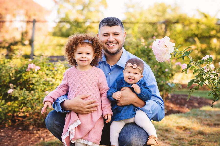 Dad poses with his 2 daughters in front of a rose garden during their family photos
