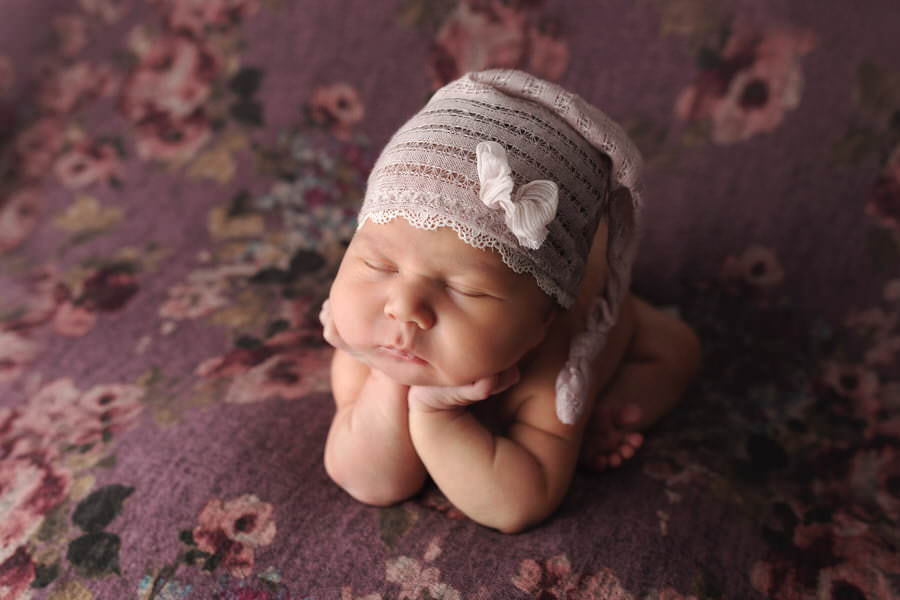 Sleeping baby girl in a pink hat posing on a purple floral blanket with her hands under her chin during her photoshoot with The Flash Lady