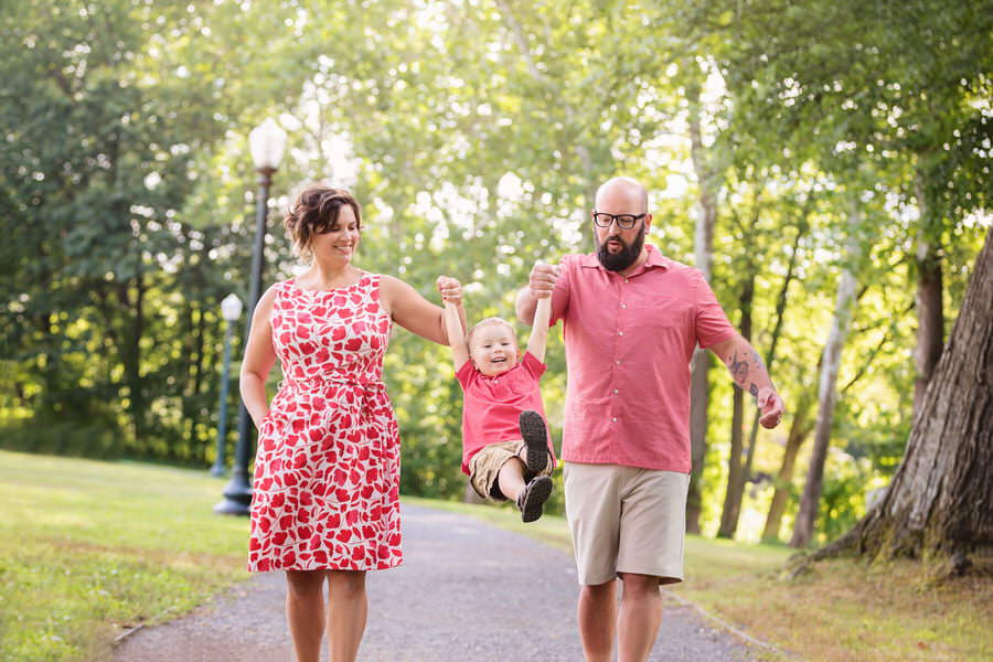 Family of 3 dressed in red swing their toddler son while walking down a path during their family photoshoot