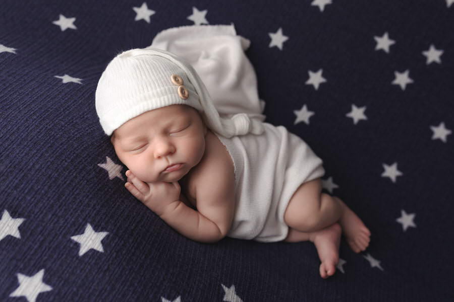 Sleeping newborn baby boy in a white wrap and hat posing on a navy and white star blanket during his photoshoot with The Flash Lady