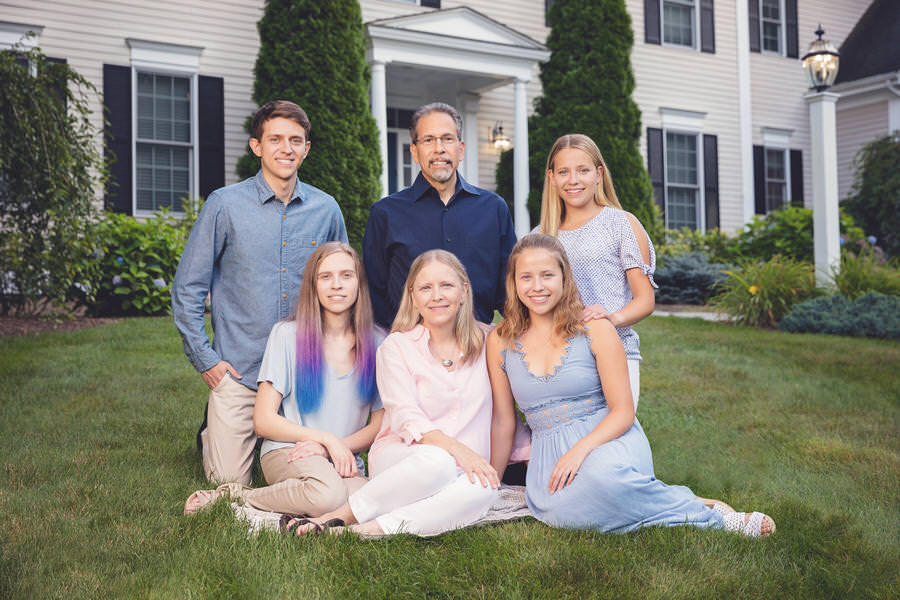 2 parents with 4 young adult children posing in blue outfits in front of their home during their family portrait session