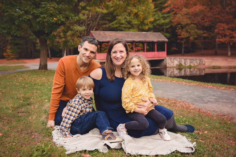 Mom and Dad sitting with their twin son and daughter on a blanket in front of a red covered bridge during their family photoshoot