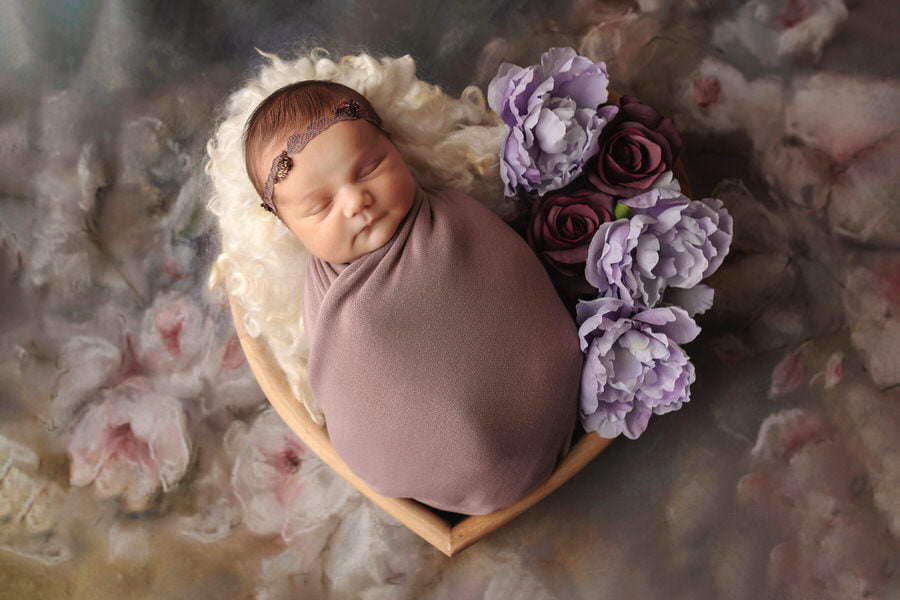 Sleeping newborn baby girl in a purple wrap and headband in a heart bowl with purple flowers during her photoshoot with The Flash Lady
