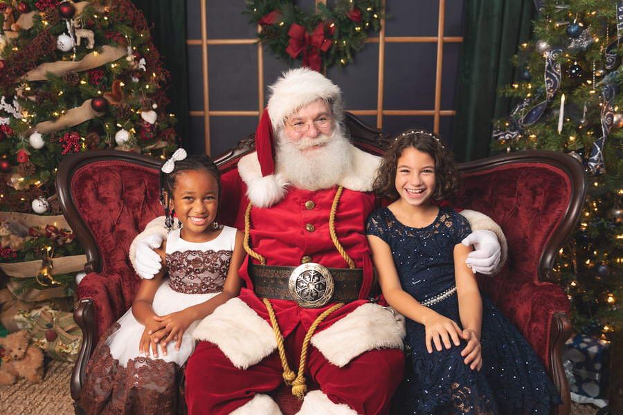 Santa Claus sitting on a red couch smiling with 2 young girls during their Christmas photoshoot