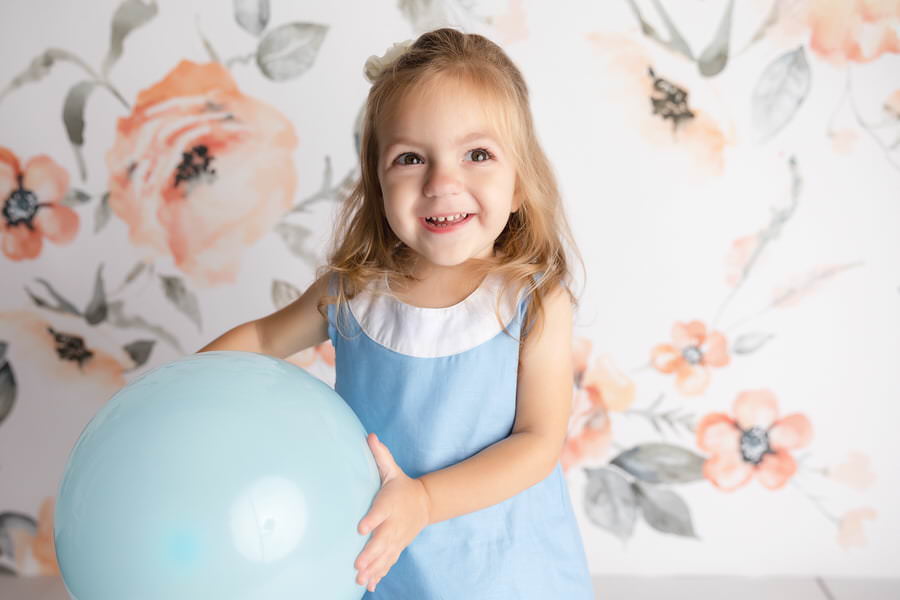 Smiling toddler girl in a blue dress holding a blue balloon during her birthday photoshoot