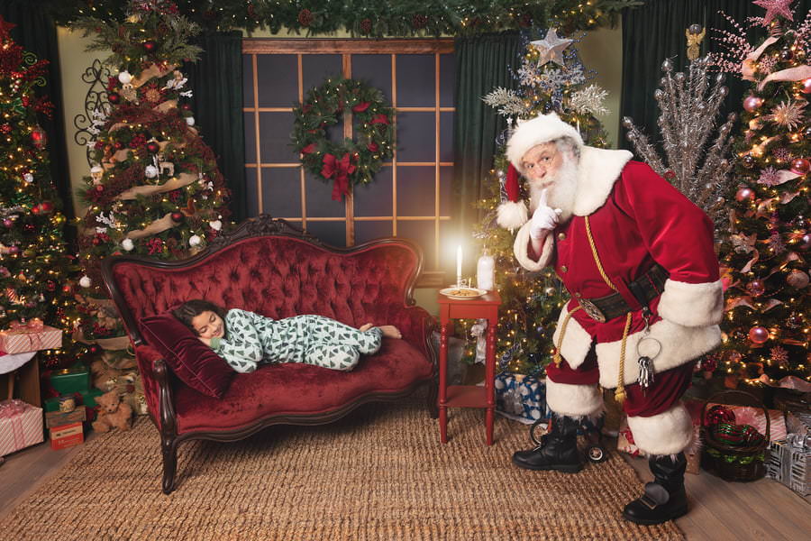 Santa telling us to Shhh as he sneaks up on a little girl sleeping on a red couch during her Photos with Santa session