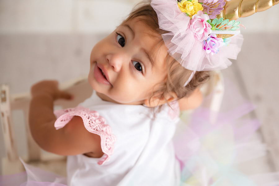 Curious baby girl in a unicorn hat turned around looking at the camera during her first birthday photoshoot