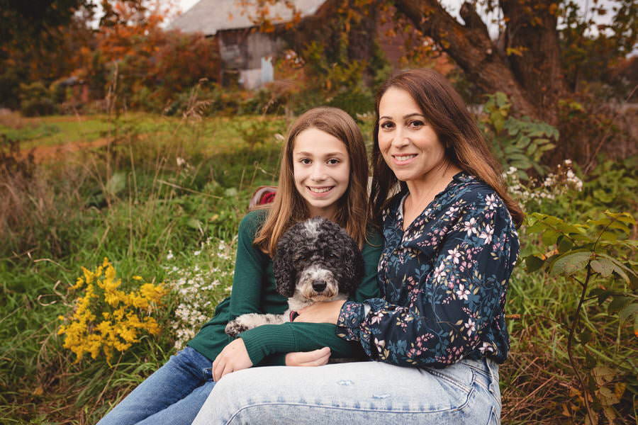 Mom sits in a red chair with her daughter and their dog in front of a barn during their family photoshoot