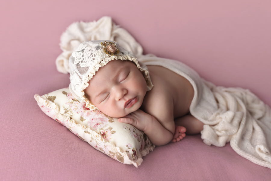 Sleeping baby girl posing on a pink blanket lying on a floral pillow in a white bonnet during her photoshoot with The Flash Lady