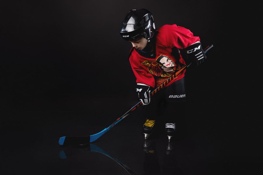 Toddler boy in a red hockey uniform holding his stick preparing to hit the puck during his birthday photoshoot