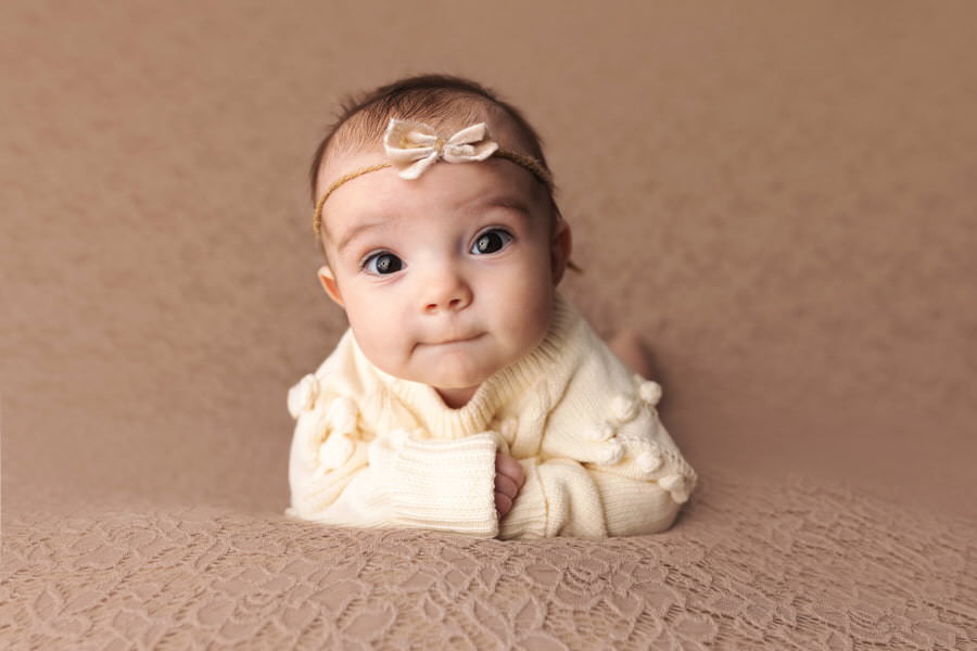 Alert 3 month old baby girl lying on her stomach in a cream outfit posing for her baby photoshoot with The Flash Lady Photography