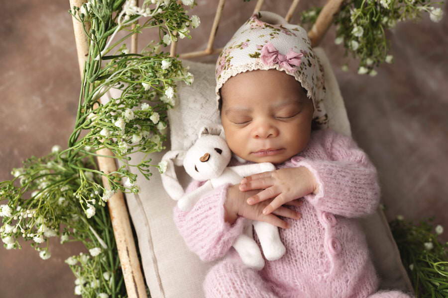Sleeping black newborn girl in pink pajamas and floral hat posing with a white bunny in a bed during her photoshoot with The Flash Lady