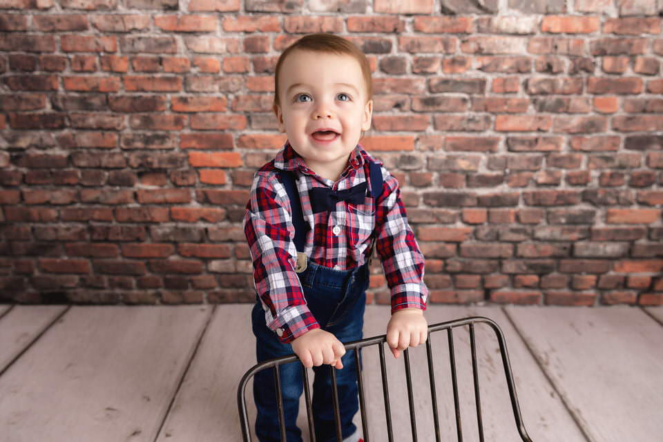 Laughing baby boy in a red plaid shirt and bow tie standing in front of a brick backdrop holding a chair during his cake smash photoshoot