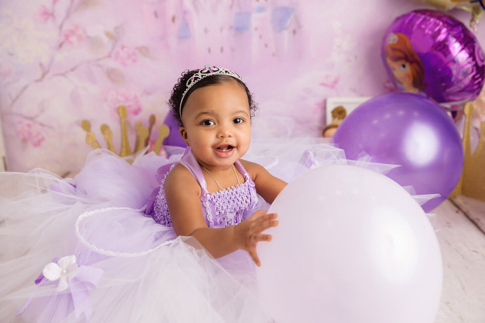 Smiling baby girl in a purple tulle dress holding a balloon during her first birthday cake smash photoshoot