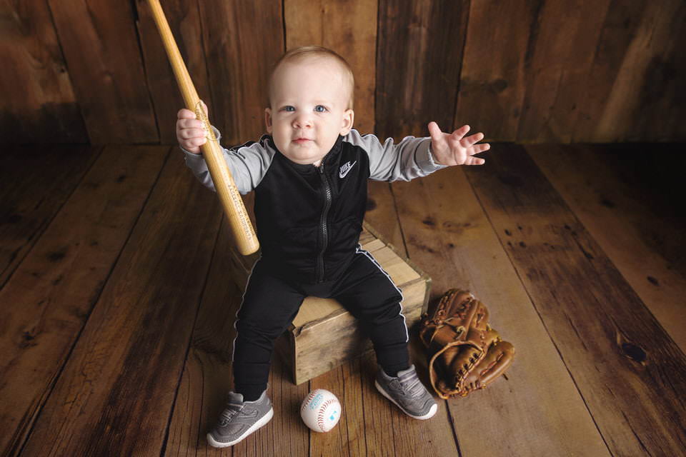 Baby boy in a black and gray tracksuit holds a baseball bat sitting on a crate during his birthday photoshoot