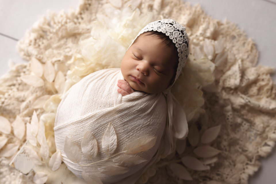 Sleeping hispanic baby girl posing in a cream wrap and bonnet on a white fur with leaves during her photoshoot with The Flash Lady