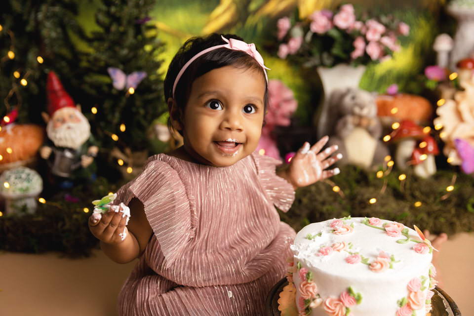 Smiling baby girl eating cake in front of her fairy garden cake smash scene
