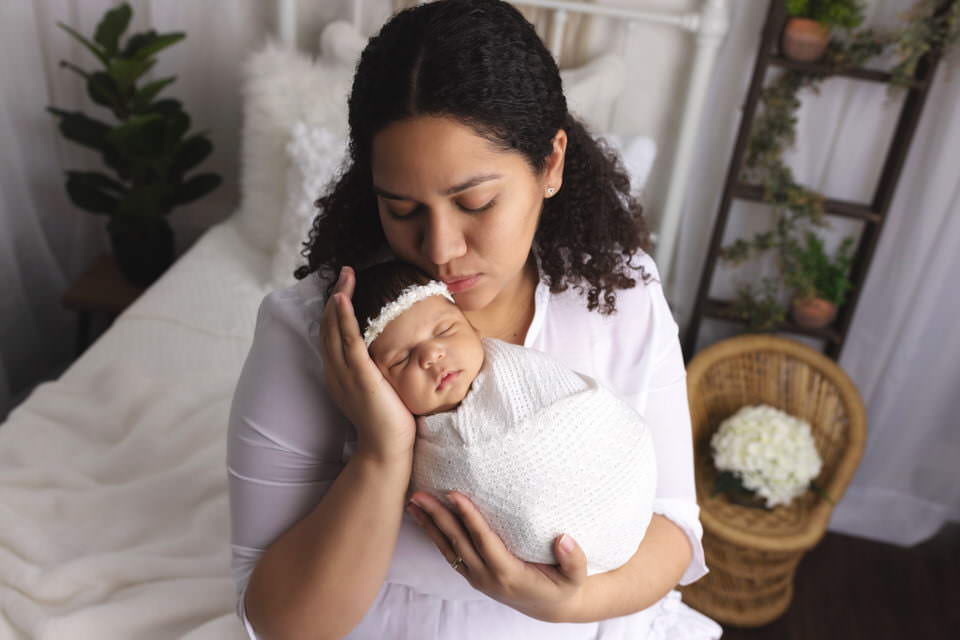 Hispanic Mom holding her baby girl wrapped in while kissing her on the head white they sit on a white bed during their photoshoot with The Flash Lady