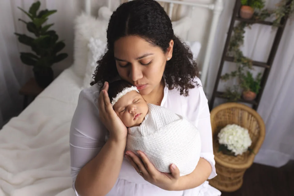 Hispanic Mom holding her baby girl wrapped in while kissing her on the head white they sit on a white bed during their newborn photoshoot