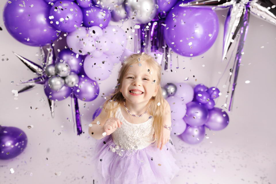 Laughing toddler girl anticipating silver confetti hitting her face standing in front of purple balloons during her birthday photoshoot with The Flash Lady Photography