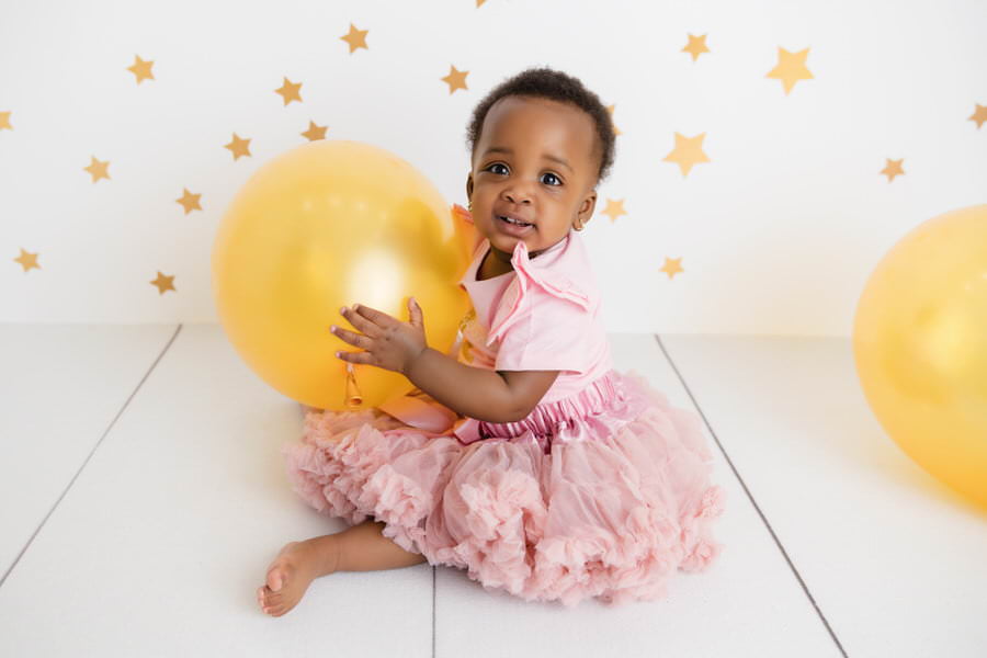 Smiling black baby girl poses in a pink outfit with gold balloon during her cake smash photoshoot with The Flash Lady Photography