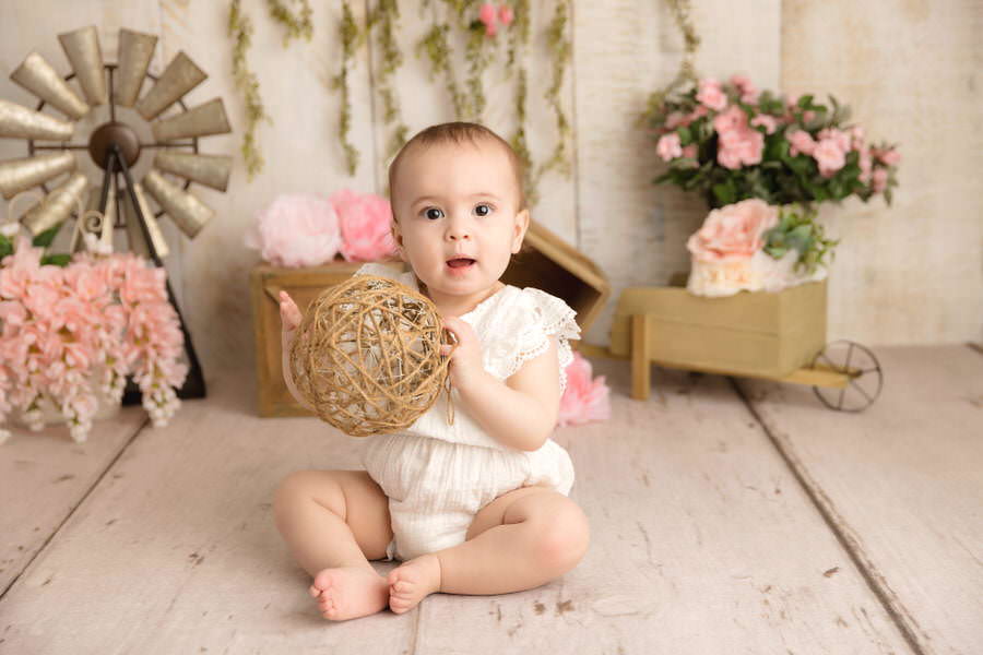 Smiling baby girl holding a rope ball in a cream romper during her first birthday cake smash photoshoot