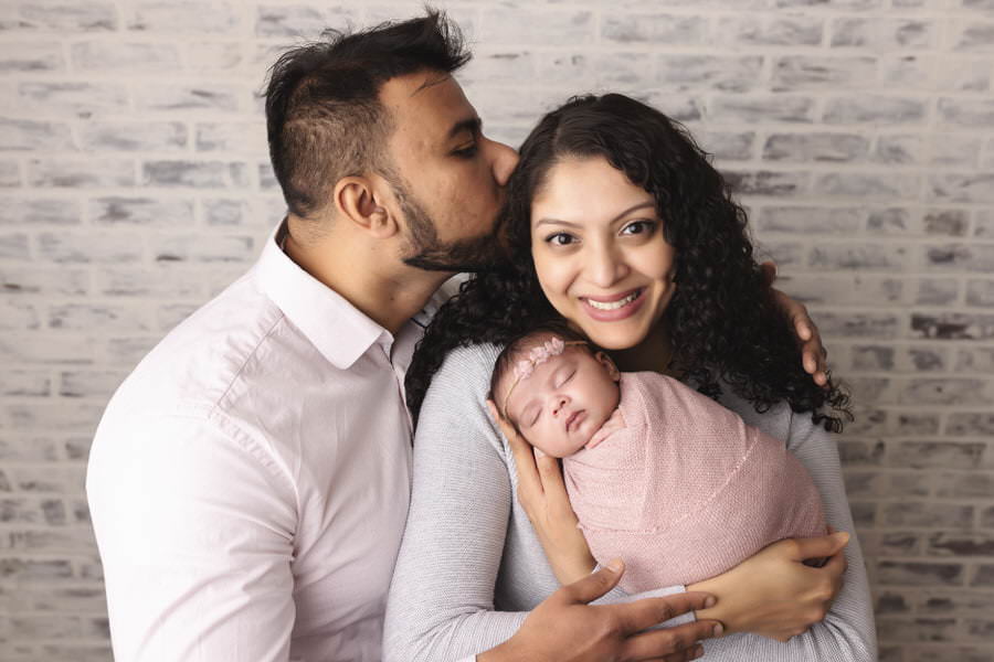 Smiling Mom posing with her newborn baby wrapped in pink as her husband kisses her head during their photoshoot with The Flash Lady