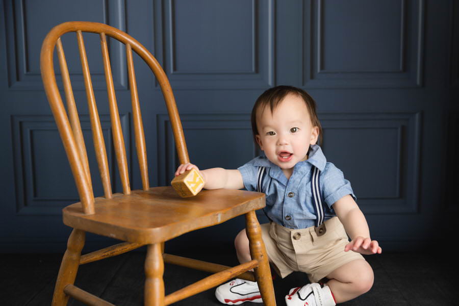 Smiling korean baby boy sitting next to a chair holding a block during his first birthday photoshoot
