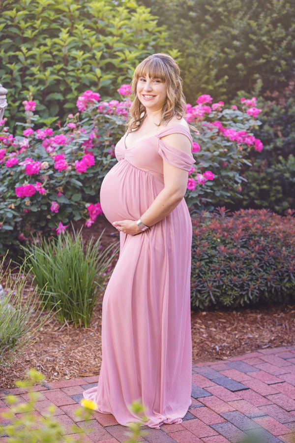 Smiling Mom posing next to pink roses during her outdoor maternity photoshoot