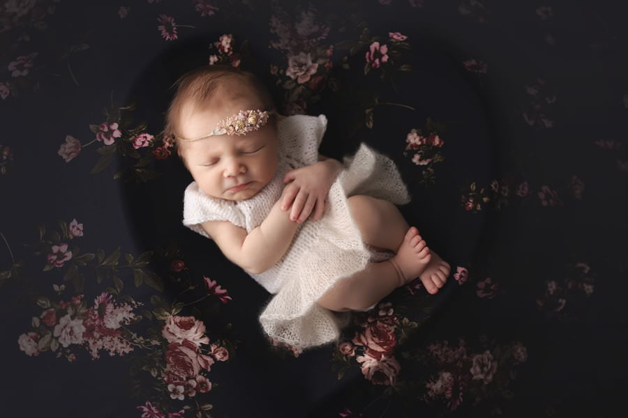 Sleeping newborn girl in a white knitted dress posing in a heart bowl on a navy blue floral blanket during her photoshoot with The Flash Lady