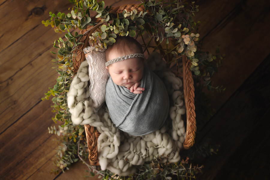 Sleeping baby girl in a sage green wrap posing in a chair with greenery surrounding her during her photoshoot with The Flash Lady