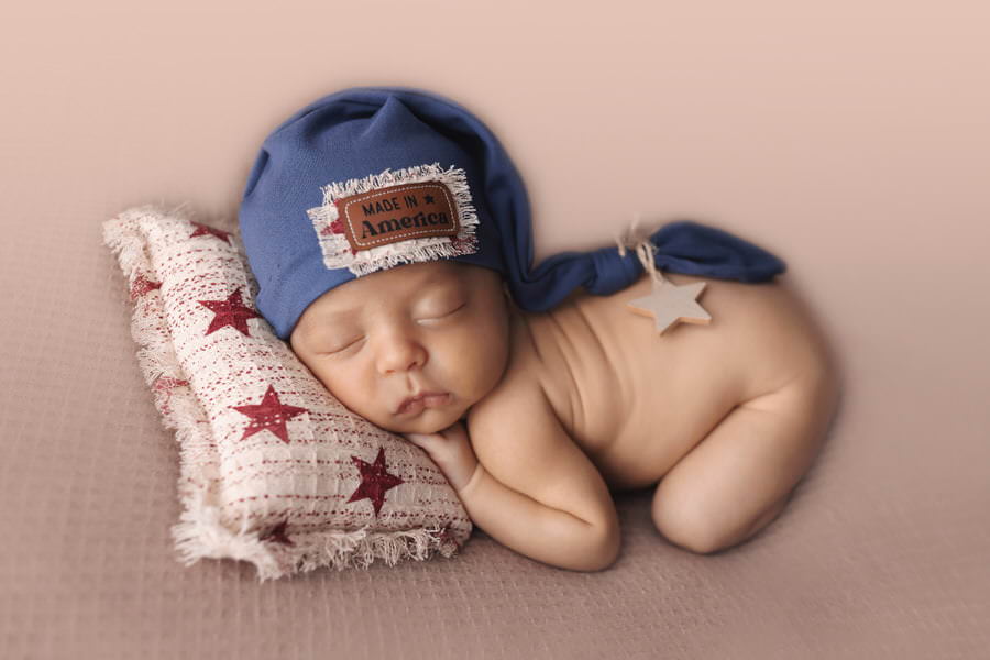 Sleeping newborn baby in a blue hat on a red and white star pillow on a tan blanket during his photoshoot with The Flash Lady