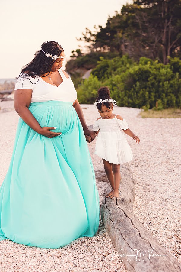 Pregnant Mom in a turquoise dress guiding her toddler daughter on a piece of driftwood