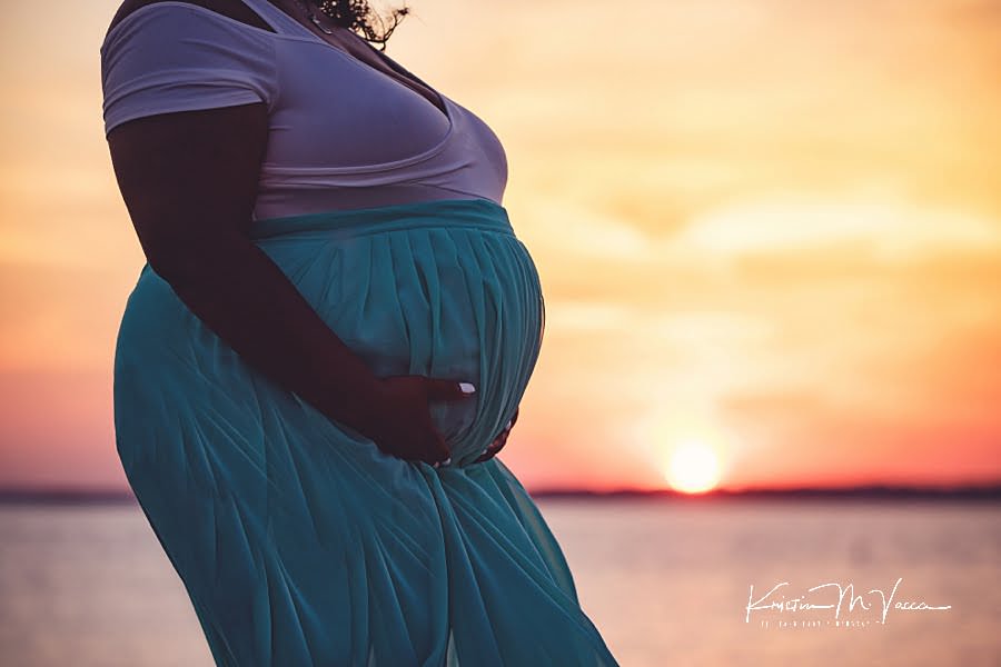 Silhouetted photo of pregnant belly in a turquoise dress against a setting sun