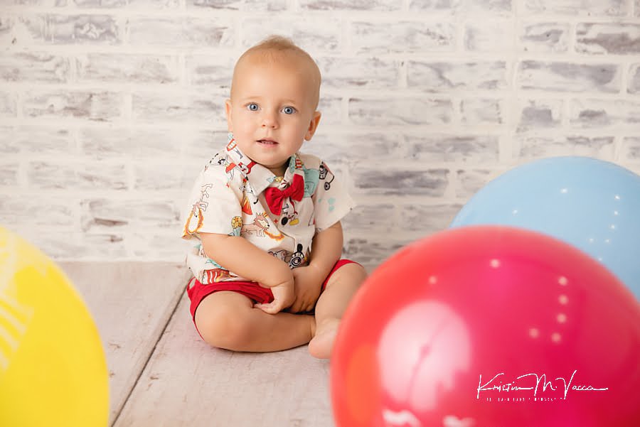 Baby boy sitting in front of a white brick wall surrounded by balloons during his first birthday photoshoot
