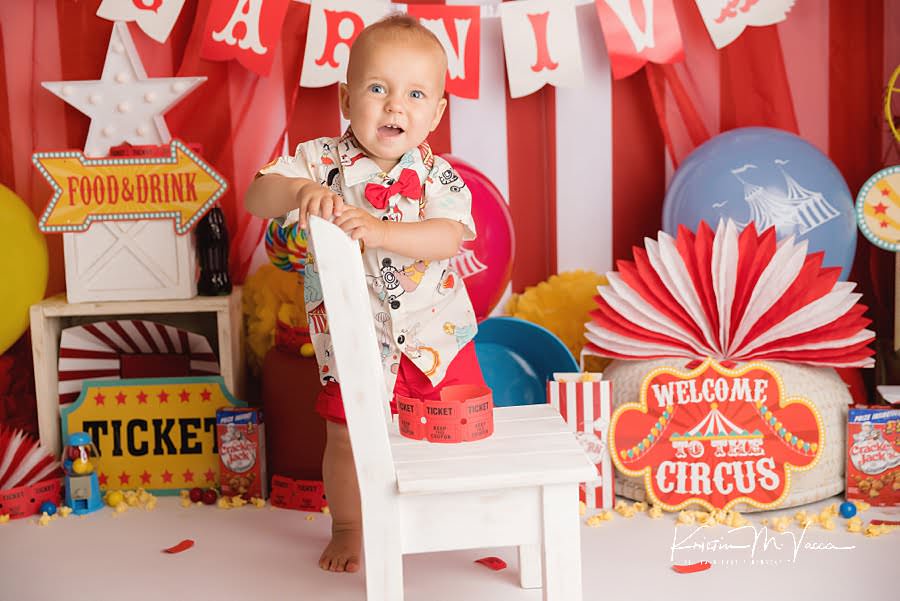 Baby boy standing behind a white chair in front of his colorful cake smash photo scene