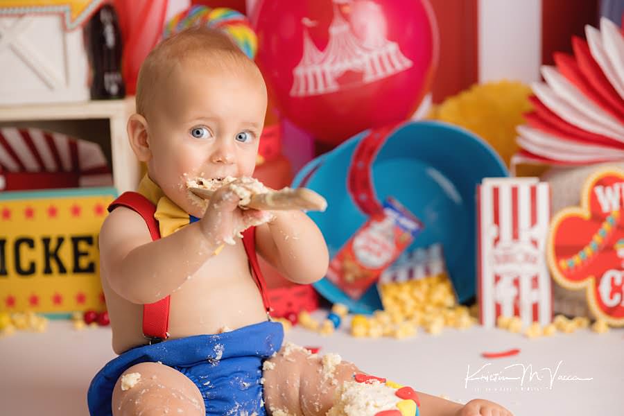 Baby boy eating cake off of a wooden spoon during his colorful carnival cake smash