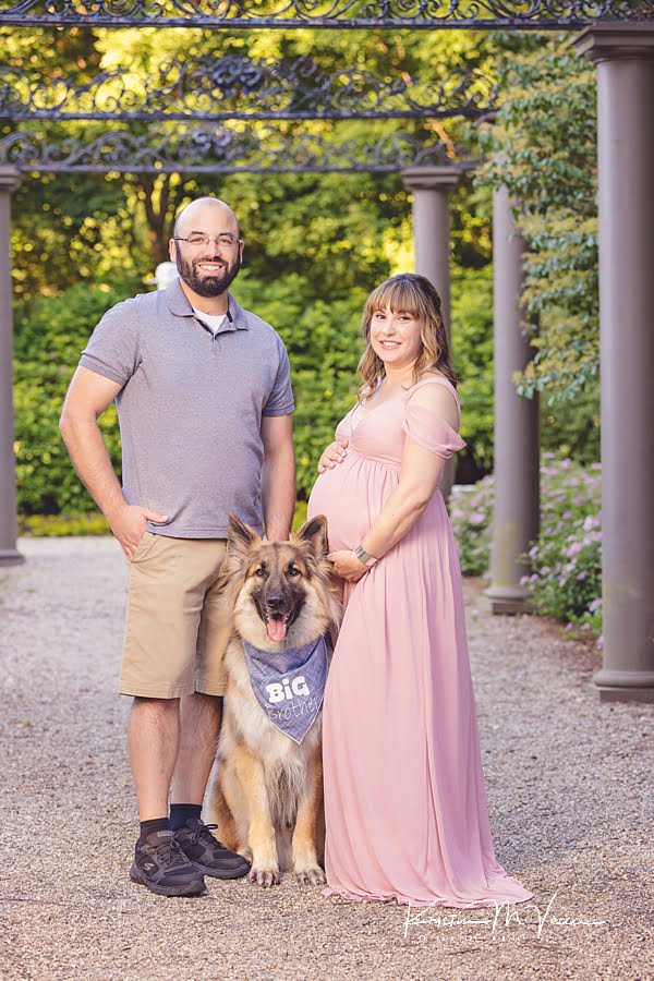 Husband and wife standing in front of pillars and greenery with their dog during their family and dog maternity photos