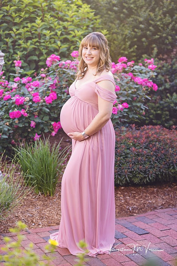Smiling pregnant woman standing in front of pink roses posing during her maternity photoshoot