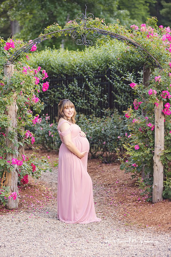Smiling pregnant woman posing for the camera standing in front of pink roses during her maternity photoshoot
