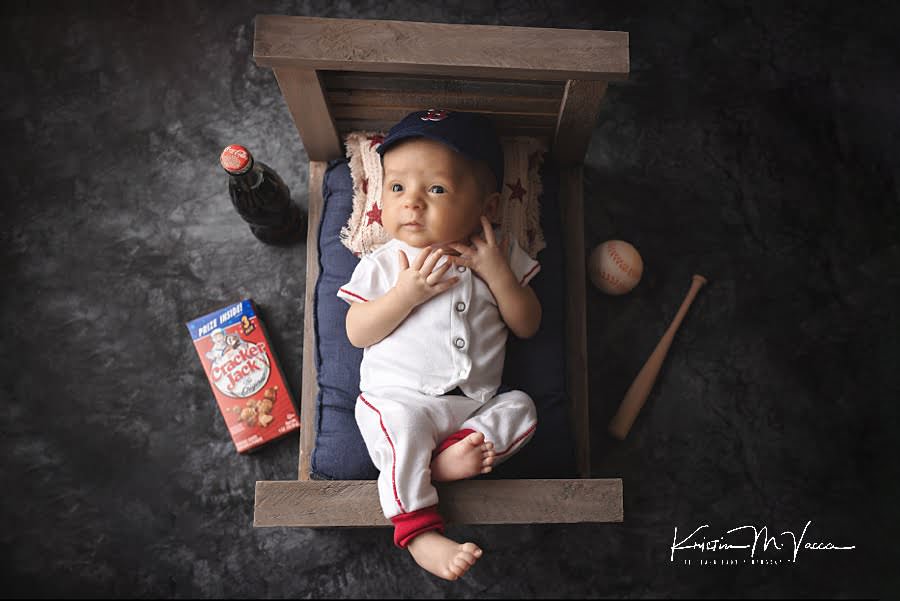 Awake infant boy in a baseball uniform posing on a bed with baseball props during his photoshoot