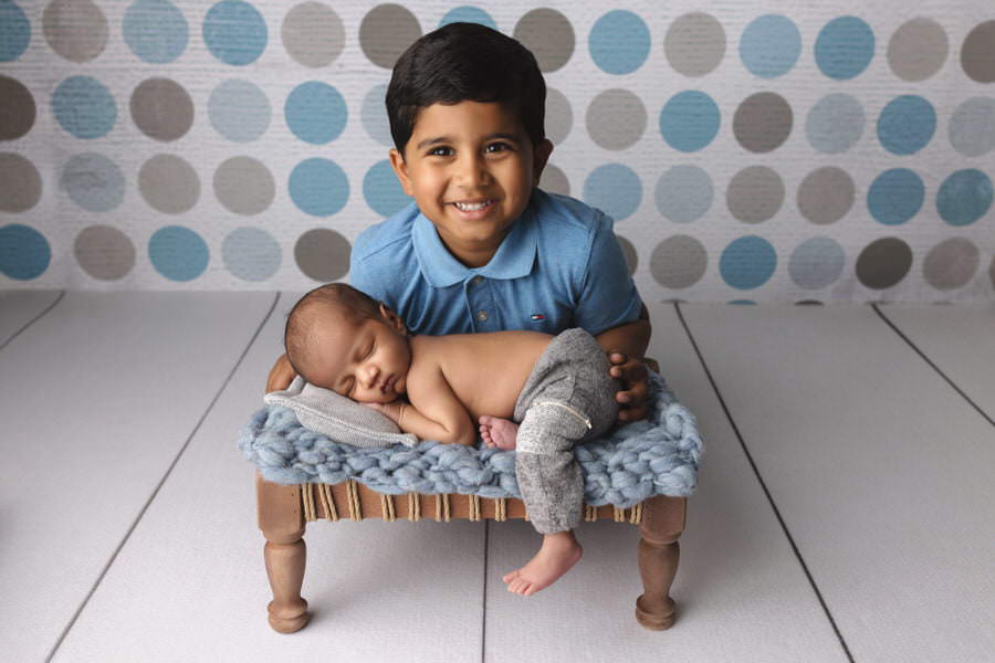 Indian newborn baby boy sleeping on a blue blanket and brown platform while his big brother smiles at the camera during their newborn photos with older brother by The Flash Lady