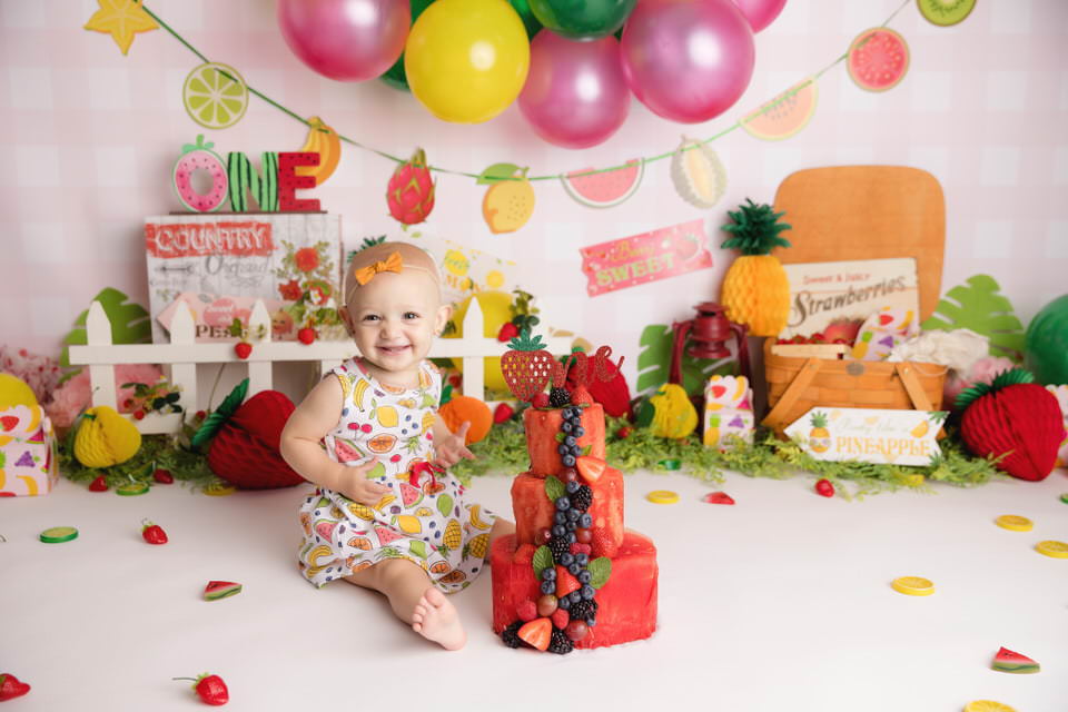 Smiling baby girl in a fruit patterned dress gets ready to smash her fruit cake during her birthday fruit themed cake smash photoshoot