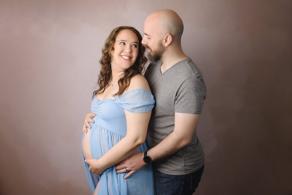 Smiling woman in a blue dress gazes back at her husband while holding her belly during her maternity photoshoot
